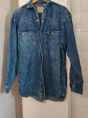 Klassisk vintage jeansskjorta strl L - Snygg blå jeansjacka i 100% bomull med två stora bröstfickor och knappar framtill. En av tryckknapparnas baksida missas, men inget som syns (mitt på)
