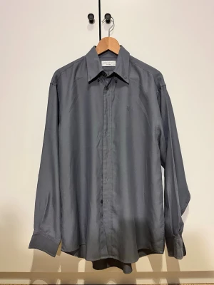 Grå skjorta från Yves Saint Laurent - Snygg grå skjorta från Yves Saint Laurent i väldigt bra skick.  Storlek Medium men den passar mer som Large (för stor för mig som är 183).