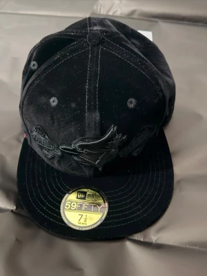 Svart keps New Era 59FIFTY Blue Jays - Snygg svart keps från New Era 59FIFTY med Blue Jays-logga broderad framtill. Kepsen har en klassisk rak skärm och är tillverkad i sammet för en lyxig känsla. Perfekt för dig som vill sticka ut med stilren accessoar.