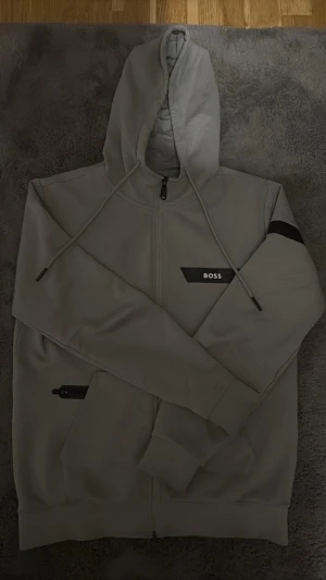 Boss zip - Snygg grå hoodie från BOSS med dragkedja framtill, huva med dragsko och svarta detaljer. Märkeslogga på bröstet och en svart detalj på ena ärmen. Perfekt för en avslappnad och stilren look.