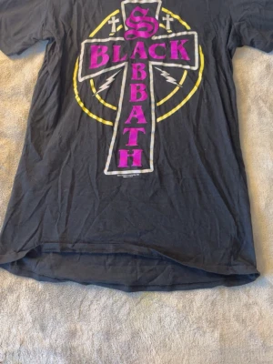 Black Sabbath t-shirt med tryck - Svart t-shirt med stort Black Sabbath-tryck i lila och gult framtill. Motiv med kors och blixtar, klassisk bandstil. Perfekt för dig som gillar rock och vill sticka ut med en ikonisk bandtröja.