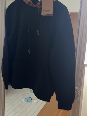 Svart hoodie från Burberry - Snygg svart hoodie från Burberry med klassisk rutig huva i beige, svart och rött. Hoodien har dragsko vid huvan och ribbade muddar vid ärmslut och nederkant. Perfekt för en trendig och avslappnad stil.