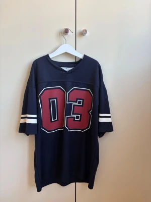 Oversized mörkblå t-shirt med tryck - Snygg oversized mörkblå t-shirt med stort rött 03-tryck framtill och vita ränder på ärmarna. Perfekt för en sportig och avslappnad stil. T-shirten har v-ringad hals och korta ärmar.