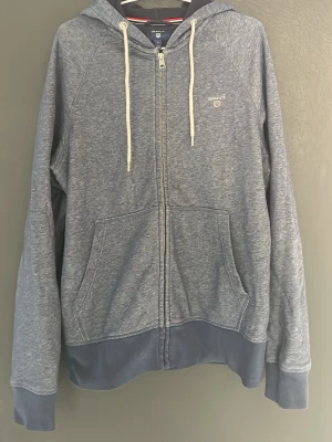 Gant tröja - Mörkgrå/blå hoodie med dragkedja från Gant. Tröjan har huva med vita snören, två stora fickor framtill och ribbade muddar. Liten Gant-logga broderad på bröstet. Perfekt för en avslappnad och stilren look.