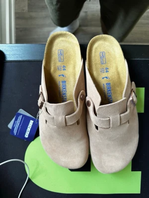 Beige Birkenstock Boston tofflor - Säljer ett par beige Birkenstock Boston tofflor i mocka med justerbart spänne och klassisk korksula. Perfekta för dig som gillar stilrena och bekväma skor. Modellen har en öppen häl och mjuk innersula för extra komfort.