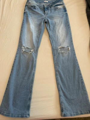 Ljusblå bootcut jeans med slitningar - Säljer ett par ljusblå bootcut jeans med slitna hål på båda knäna. Jeansen har klassisk femficksdesign och är tillverkade i denim. Snygga och sköna. 