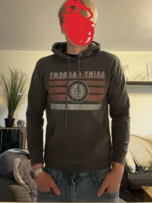 Mörkgrå Saint Laurent hoodie - Snygg mörkgrå hoodie från Saint Laurent med tryck framtill i rött, vitt och grått. Klassisk huva med dragsko och stor magficka. Perfekt för en avslappnad och trendig stil.