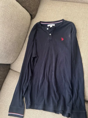 Mörkblå långärmad topp U.S. Polo Assn. - Mörkblå långärmad topp från U.S. Polo Assn. med två knappar vid halsen och röd broderad logga på bröstet. Tillverkad i mjuk bomull och har en klassisk, avslappnad passform. Perfekt för en stilren och enkel look. Finns fläck som ni ser