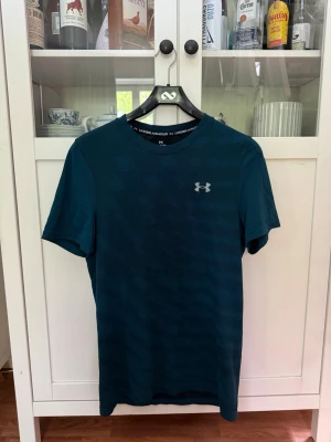 Mörkgrön träningströja från Under Armour - Snygg mörkgrön t-shirt från Under Armour med diskret mönster och logga på bröstet. Tröjan har rund halsringning och är perfekt för träning och sport.