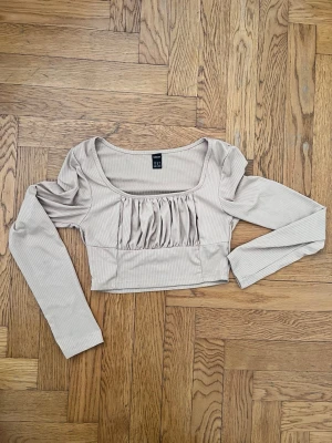 Beige ribbad topp  - Trendig beige ribbad topp  med långa ärmar och croppad passform. Toppen har rynkade detaljer framtill och en fyrkantig urringning som ger en snygg siluett. Perfekt att styla med jeans eller kjol för en cool look.