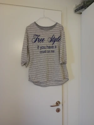 Randig grå topp med text - Grå och vit randig topp med trekvartsärm och rund halsringning. Tryck i blått framtill med texten 'Free Style if you have a crush on me'. Skön och avslappnad passform, perfekt till jeans eller leggings.
