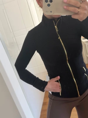 Svart träningsjacka från Lululemon black gold svart guld S stl 2  - Snygg svart träningsjacka från Lululemon med guldiga dragkedjor och figurnära passform. Jackan har hög krage, långa ärmar och två fickor framtill med dragkedja. Perfekt för gymmet eller löprundan. Storlek 2 passar S 