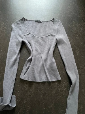 Grå ribbad långärmad topp - Snygg grå ribbad topp från Gina tricot med lång ärm och unik v-ringad skärning vid halsen. Toppen har en figurnära passform och är tillverkad i ett mjukt, stretchigt material som sitter bekvämt. Perfekt att styla med jeans eller kjol.