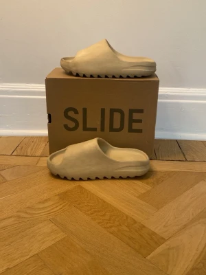 Beige Adidas Yeezy Slide tofflor - Säljer ett par beige Adidas Yeezy Slide tofflor med chunky sula och öppen tå. Tillverkade i mjukt och flexibelt skum för maximal komfort. Perfekta för chill dagar hemma eller när du vill ha en clean och trendig look.