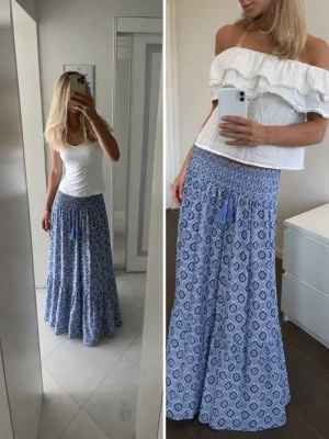 Blå mönstrad långkjol med tofsar - Snygg blå långkjol med vitt och mörkblått mönster. Kjolen har smock i midjan och dekorativa tofsar framtill. Den är luftig och perfekt för varma dagar. Passar till både enkla toppar och mer dressade looks.