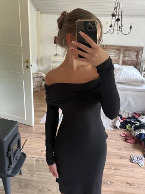 Svart offshoulder klänning - Snygg svart klänning med offshoulder-design och långa ärmar. Klänningen har en tight passform och är tillverkad i ett stretchigt material som sitter skönt på kroppen. Perfekt för dig som gillar en stilren och elegant look.