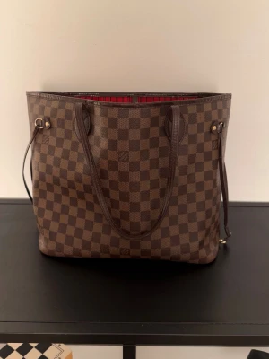 Louis Vuitton Damier Neverfull MM - Snygg shoppingväska från Louis Vuitton i klassiskt brunt Damier Ebene-mönster. Väskan har långa handtag, rödrandigt innerfoder och detaljer i mörkbrunt läder. Perfekt för dig som vill ha en rymlig och trendig väska med lyxig känsla.