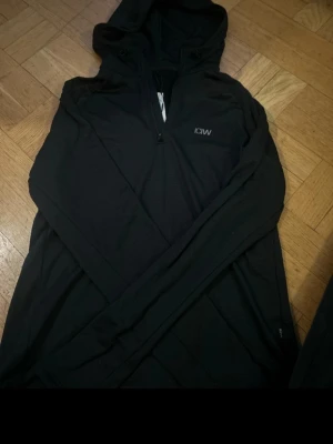 Svart hoodie från ICANIWILL - Svart långärmad hoodie från ICANIWILL med halv dragkedja framtill och diskret logga på bröstet. Tillverkad i ett lätt och stretchigt funktionsmaterial som passar perfekt till träning eller löpning. Huva för extra komfort och stil.