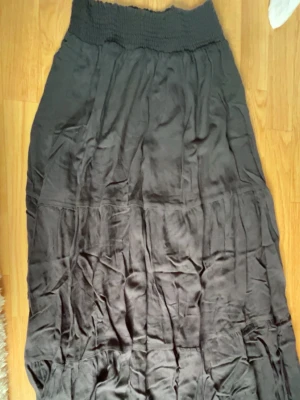 Svart långkjol från 157, smockad midja - Svart långkjol från 157 med smockad, elastisk midja. Kjolen har en flowy passform och är tillverkad i ett mjukt tyg som ger en avslappnad look. Perfekt att styla med en crop top eller stickad tröja.