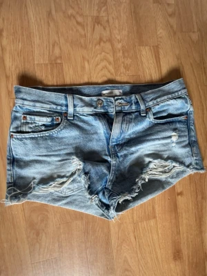 Ljusblå denimshorts med slitningar - Snygga ljusblå denimshorts från Gina Tricot med råa kanter och slitna detaljer framtill. Klassisk femficksmodell med bälteshällor och knappgylf. Perfekta för varma dagar och en avslappnad stil.
