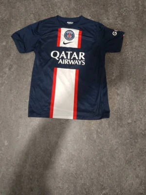 PSG matchtröja blå Nike - Snygg blå PSG matchtröja Nike med Qatar Airways-tryck på bröstet. Tröjan har vita och röda vertikala ränder framtill, klubbmärke och Nike-logga. Tillverkad i lätt och ventilerande material, perfekt för fotboll eller träning.