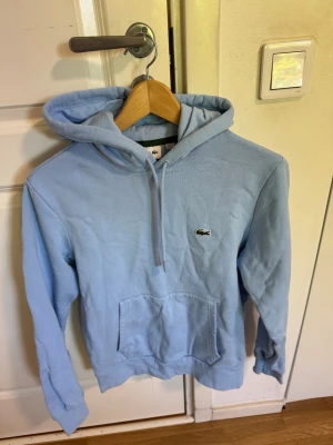 Ljusblå hoodie från Lacoste - Snygg ljusblå hoodie från Lacoste med klassisk passform. Hoodien har huva med dragsko, stor magficka och den ikoniska krokodilloggan broderad på bröstet. Tillverkad i mjukt bomullsmaterial som känns skönt mot huden. Perfekt för en chill och avslappnad stil.