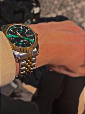 Grön och guld klocka Keep Moving - Stilren klocka från Keep Moving med grön urtavla, guldfärgade detaljer och romerska siffror. Armbandet är i tvåfärgad metall, silver och guld, och boetten har en räfflad guldkant. Perfekt accessoar för att lyfta din outfit.