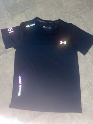 Svart träningströja från Under Armour - Svart kortärmad träningströja från Under Armour med reflekterande logga på bröstet och text på ärmen samt sidan. Tillverkad i ett lätt och snabbtorkande funktionsmaterial, perfekt för intensiva träningspass.