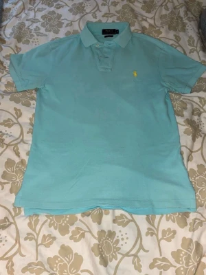 Ljusblå pikétröja från Polo Ralph Lauren - Snygg ljusblå pikétröja från Polo Ralph Lauren med klassisk krage och korta ärmar. Tröjan har den ikoniska gula loggan broderad på bröstet och är tillverkad i mjuk bomull. Perfekt för en avslappnad och stilren look.
