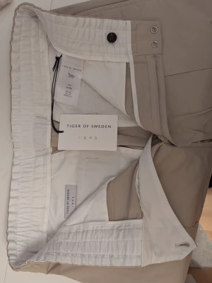 Beige chinos från Tiger of Sweden - Snygga beige chinos från Tiger of Sweden med elastisk midja och klassisk passform. Byxorna har diskreta sömmar framtill och stängs med knapp och dragkedja. Perfekta för en stilren och avslappnad look.