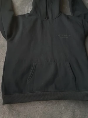 Svart Essentials Fear of God hoodie - Svart hoodie från Essentials Fear of God med broderad logga på bröstet och stor broderad text på ryggen. Klassisk känguruficka framtill och ribbade muddar. Perfekt för en clean och stilren streetwear-look.