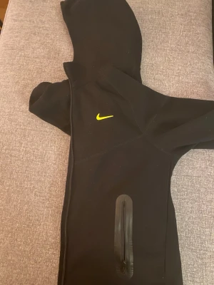 Svart Nike Nocta Kofta - Svart hoodie från Nike med gul logga på bröstet och detaljer i gult på ryggen. Hoodien har dragkedjeficka på sidan och är tillverkad i ett mjukt, stretchigt material. Perfekt för en sportig och avslappnad stil.