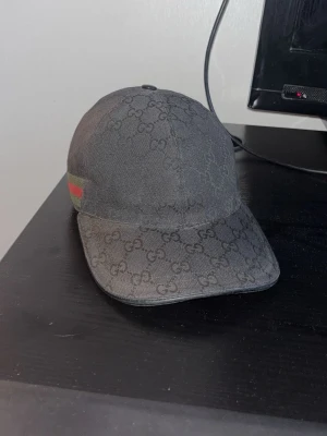 Svart Canvas Gucci keps - Svart keps från Gucci med klassiskt GG-monogram över hela kepsen och en grön-röd rand på sidan. Kepsen har böjd skärm och är tillverkad i tygmaterial. Perfekt accessoar för en trendig streetwear-look.