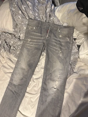 Grå slitna jeans från Dsquared2 - Säljer ett par grå slim fit jeans från Dsquared2 med slitningar och distressed detaljer. Jeansen har klassisk femficksmodell, vita färgstänk och en snygg tvättad look. Perfekta för dig som gillar streetwear och vill ha en edgy stil.