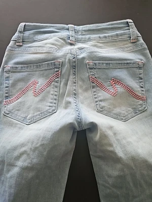 Bootcut jeans med rosa brodyr, Nelly - Ljusblå bootcut jeans från Nelly med låg midja och snygg rosa brodyr i vågigt mönster på bakfickorna. Klassisk femficksmodell i mjuk denim, perfekt för en trendig och avslappnad look.