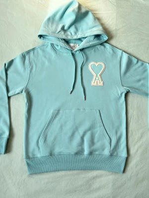 Ljusblå hoodie från Ami Paris - Snygg ljusblå hoodie från Ami Paris med stor broderad logga i form av ett hjärta och bokstaven A på bröstet. Hoodien har huva med dragsko, magficka och ribbade muddar. Perfekt för en chill och trendig look.