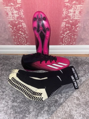 Adidas speedportal 2 (41) - DSäljer ett par snygga fotbollsskor från Adidas i rosa och svart med vita ränder på sidan. Du får ett par nya Nike pro strumpor på köper! Skorna har en låg profil, snörning och dobbar för spel på gräs. Ovandelen är i syntetmaterial för bra bollkontroll och lätt vikt.