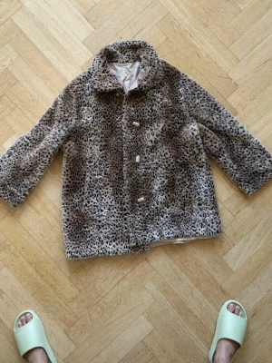 Leopardmönstrad fuskpälsjacka - Snygg leopardmönstrad fuskpälsjacka med tre knappar framtill och klassisk krage. Jackan är i beige, bruna och svarta toner och har ett mjukt innerfoder. Perfekt för dig som vill sticka ut med en trendig look under kalla dagar.