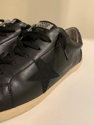 Golden Goose svarta sneakers med stjärna - Svarta sneakers från Golden Goose med ikonisk svart stjärna på sidan och glittriga detaljer på hälen. Skorna har svarta skosnören, vit sula och är tillverkade i läder. Märkeslogga syns på hälen och plösen. Perfekta för dig som gillar streetstyle.