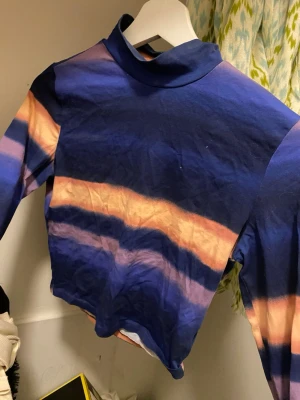Blå och orange polotopp från Monki - Säljer en cool polotopp från Monki i storlek S. Toppen har blå och orange tie-dye mönster med höghalsad krage och långa ärmar. Perfekt för dig som gillar färg och vill sticka ut lite extra.