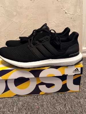 Svarta Adidas Ultra Boost sneakers - Svarta Adidas Ultra Boost sneakers med stickad ovandel och vita Boost-sulor. Skorna har svarta plastdetaljer på sidorna och hälpartiet samt klassiska tre ränder. Sitter på en färgglad originalkartong från Adidas.