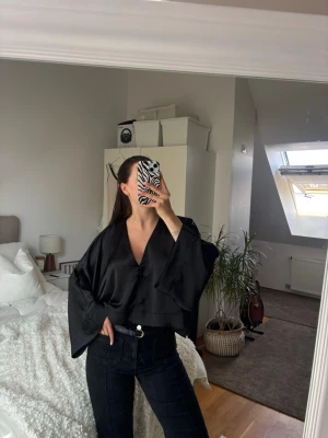 Svart blus med vida ärmar - Snygg svart satin liknande blus med djup v-ringning och vida, långa ärmar. Blusen har en croppad passform. Snyggt att ha spets under. 