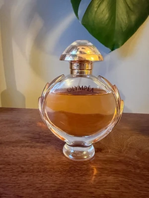 Paco Rabanne Olympea parfym - Säljer en Olympea parfym från Paco Rabanne. Flaskan är rund med detaljer i guld och har en elegant, genomskinlig design med gyllene lock. Parfymen har en varm bärnstensfärgad vätska och flaskan är tillverkad i glas.