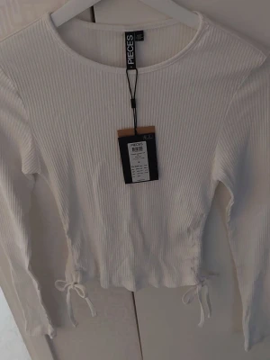 Vit ribbad croppad topp från Pieces - Snygg vit croppad topp från Pieces med långa ärmar och ribbad struktur. Toppen har knytband i sidorna för en extra detalj och sitter tajt mot kroppen. Nypris 300kr 💕