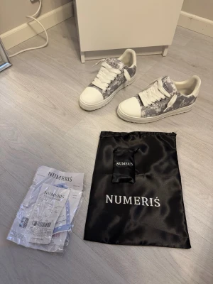 Gråvita sneakers från Numeris - Snygga sneakers från Numeris i vitt och grått med coolt mönster. Skorna har vita snören, vit sula och detaljerad logga på plösen. Perfekta för dig som gillar streetstyle och vill sticka ut med ett unikt par sneakers.