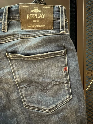 Replay anbass W 29 - Snygga blå jeans från Replay med klassisk femficksdesign och raka ben. Jeansen har en diskret tvättad look, mörkblå sömmar och en liten röd detalj på bakfickan. Märkespatch i läder baktill i midjan. Perfekta för en avslappnad stil. Storlek W 29 L 33