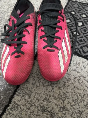 Rosa Adidas fotbollsskor med dobbar - Säljer ett par snygga fotbollsskor från Adidas i en grymt fräsch rosa färg med vita ränder på sidan och svarta detaljer. Skorna har svart plös, svarta skosnören och en ovandel i syntetmaterial med struktur för bättre bollkontroll. Perfekta för dig som vill sticka ut på planen.