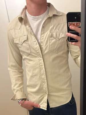 Beige skjorta från polo Ralph Lauren - Snygg beige skjorta från Polo med klassisk westernstil. Skjortan har två bröstfickor med lock och knappar, samt markerade sömmar på axlarna. Tillverkad i bomull och har lång ärm med knapp vid manschetten.
