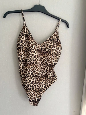 Leopardmönstrad baddräkt med smala band - Snygg baddräkt med leopardmönster och smala axelband. Djup urringning fram och klassisk skärning. Materialet är  en stretchig syntetblandning som polyester eller elastan, perfekt för strand eller pool. Aldrig använd.