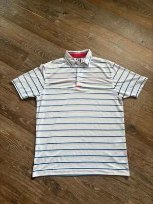 Vit randig piké från FootJoy - Snygg vit pikétröja från FootJoy med blå horisontella ränder och klassisk krage. Tröjan har korta ärmar, knappar framtill och en röd detalj vid knappslån. Perfekt för en sportig och stilren look.